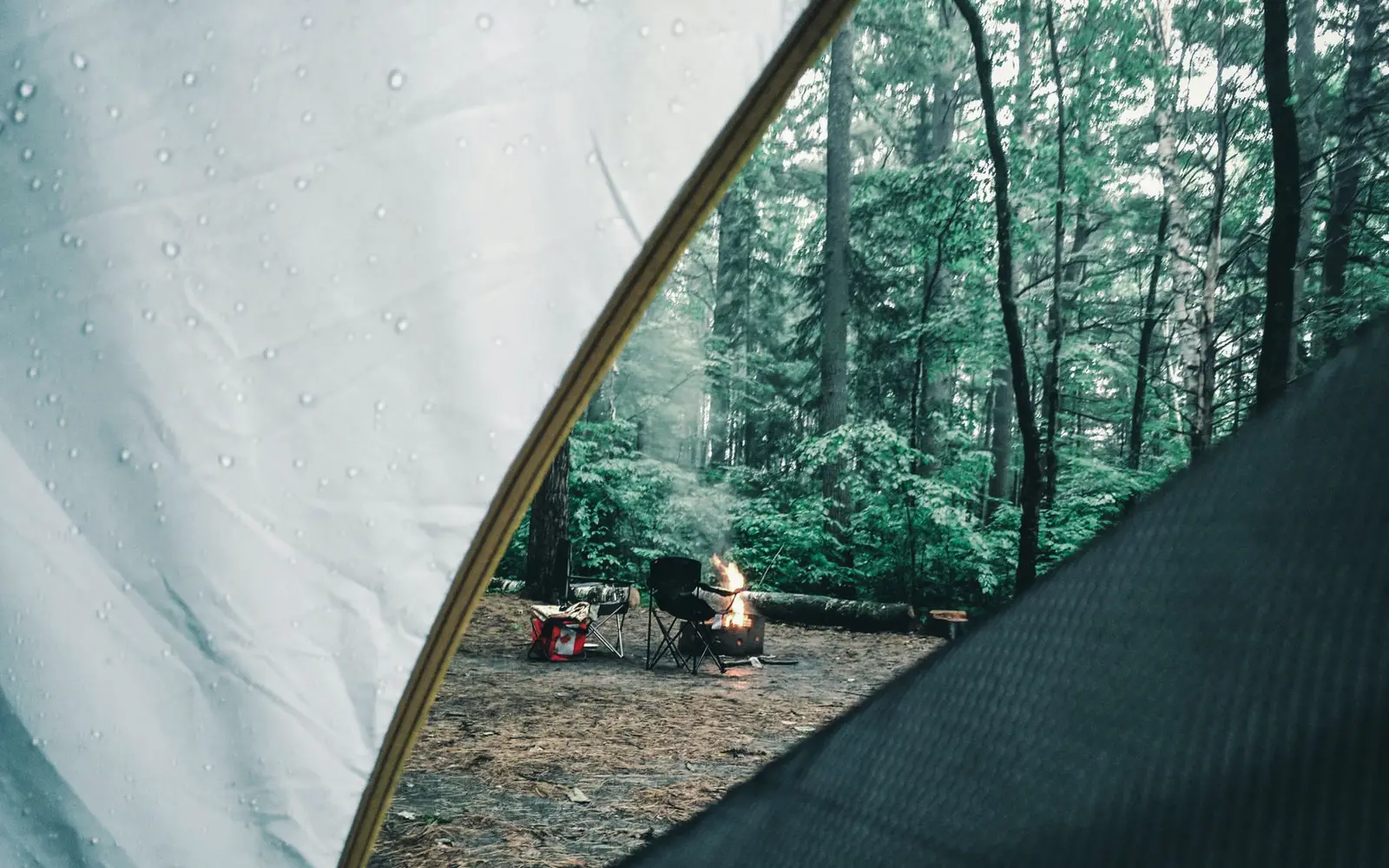 Vista desde el interior de una casa de campaña con gotas de lluvia hacia un campamento en un bosque verde