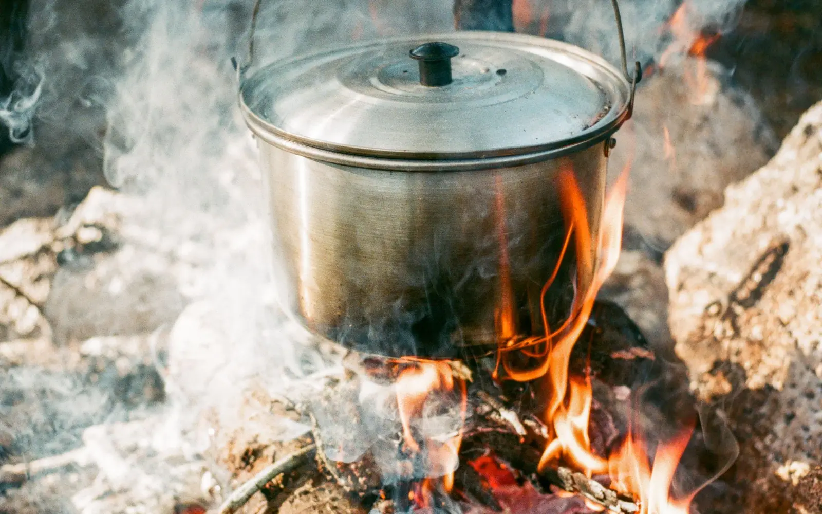 Olla de acero inoxidable con tapa calentándose sobre una fogata entre piedras y humo
