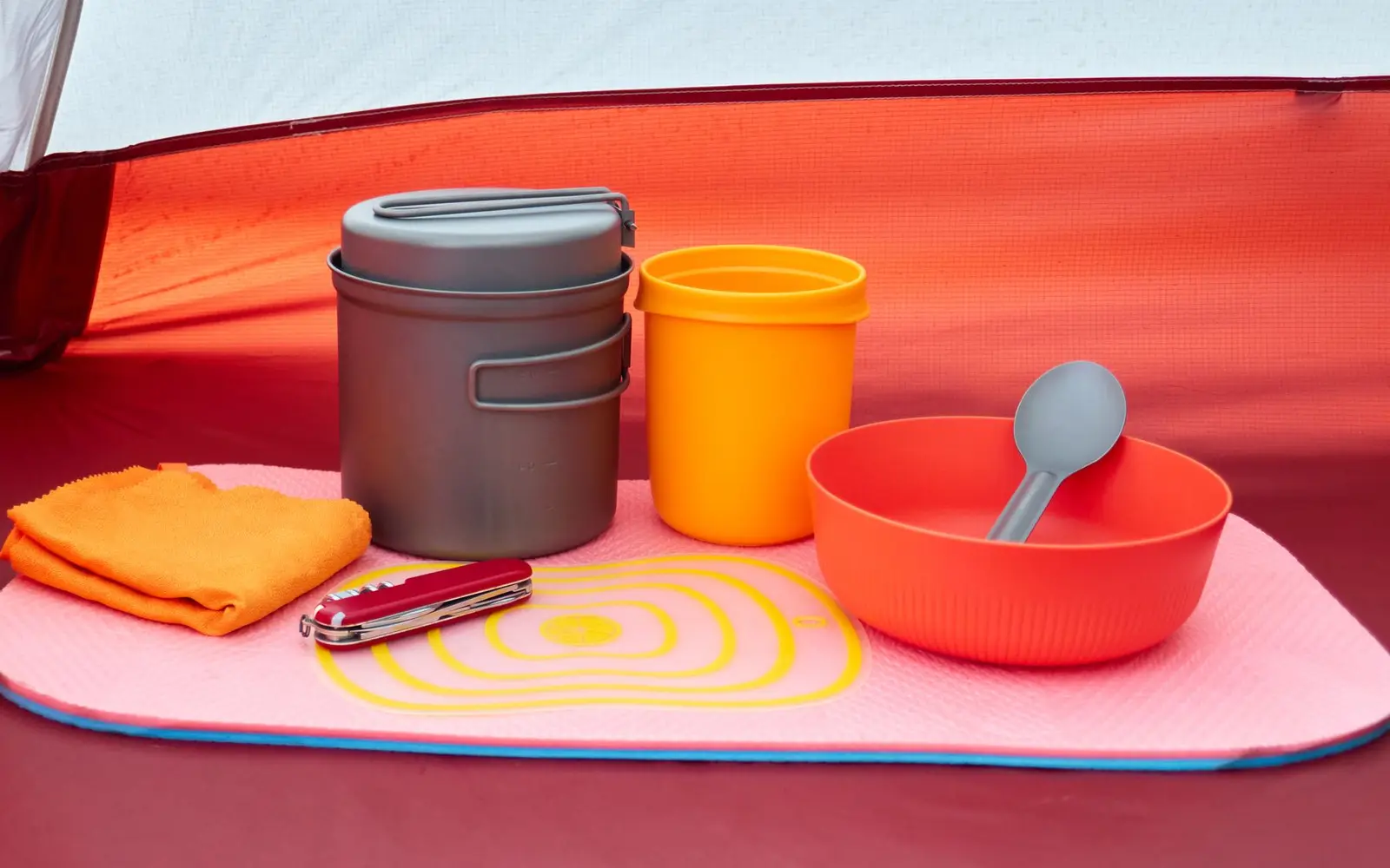 Equipo de campamento ordenado sobre un tapete: ollas de aluminio, vaso naranja, plato, cuchara y navaja dentro de una casa de campaña