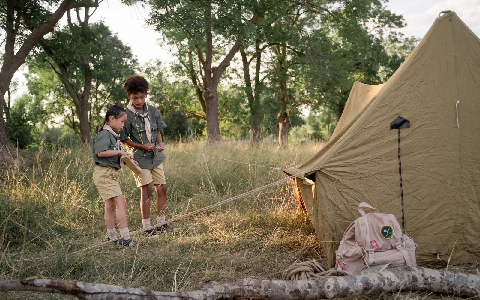 Dos niños con uniforme de exploradores armando una casa de campaña de lona en un bosque con pasto alto