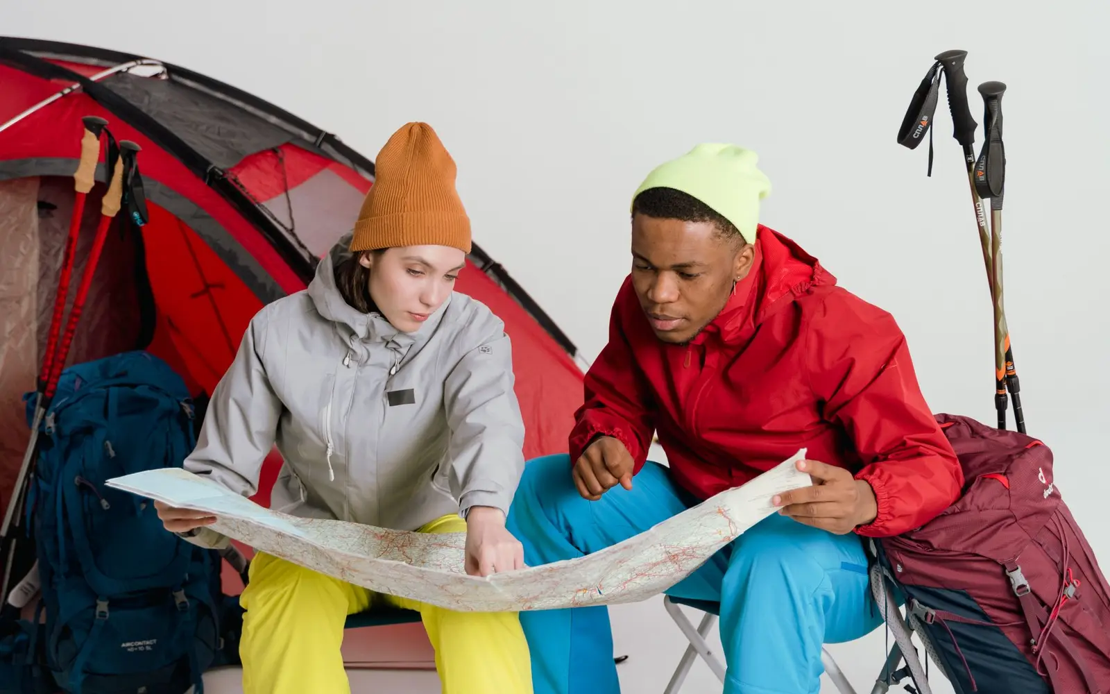 Dos campistas con gorros revisando un mapa de papel junto a una casa de campaña roja y bastones de senderismo