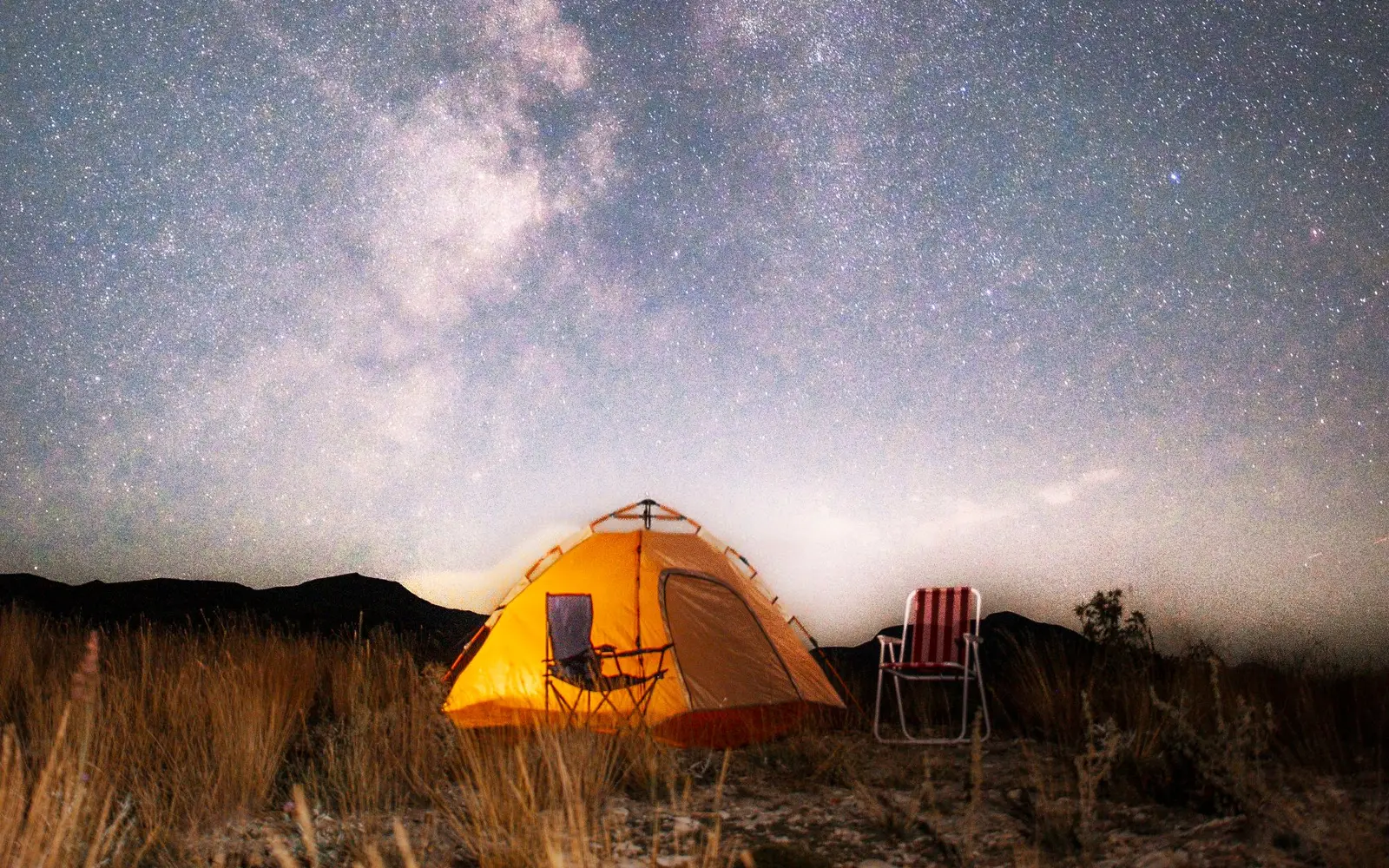 Casa de campaña naranja iluminada por dentro bajo la Vía Láctea en un cielo lleno de estrellas