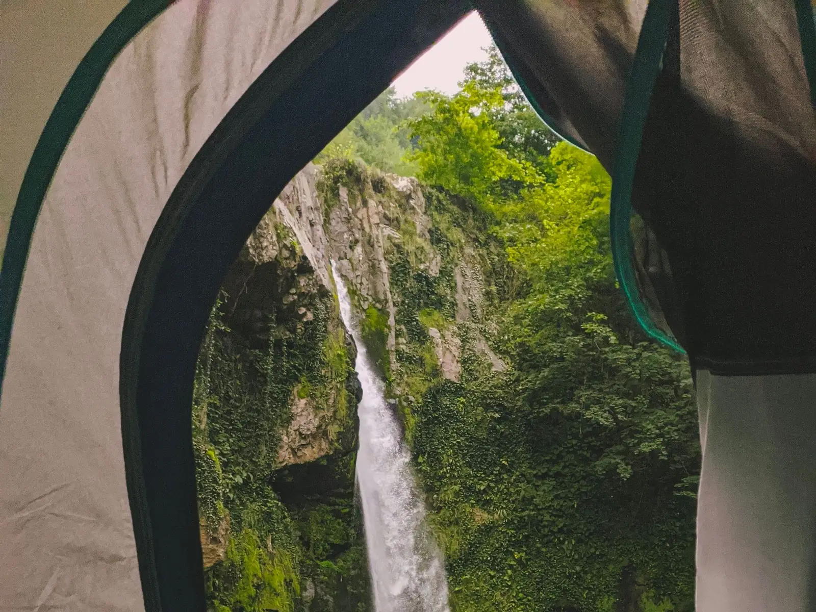Vista de una cascada entre vegetación verde desde la entrada abierta de una casa de campaña